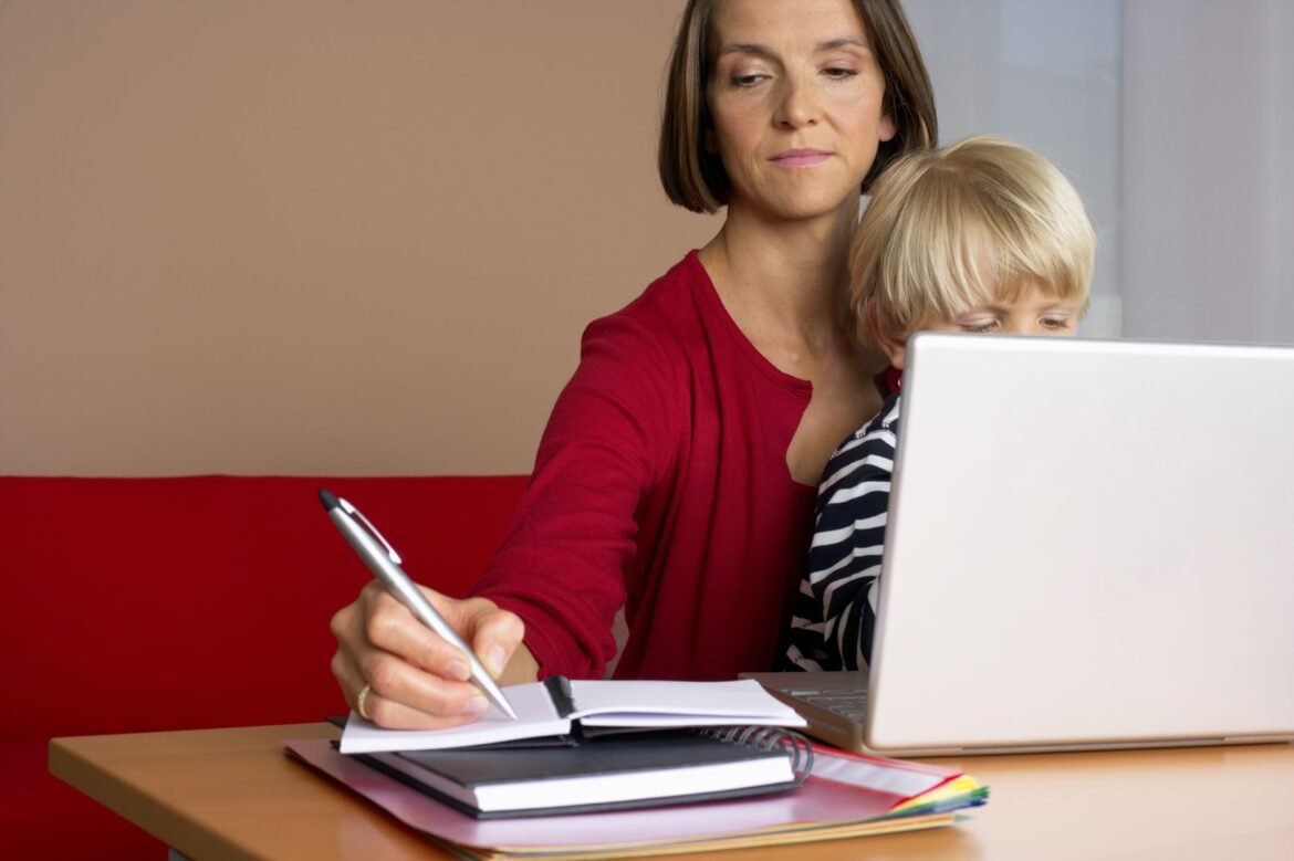 A woman multitasks with a child on her lap while working on a laptop at home.